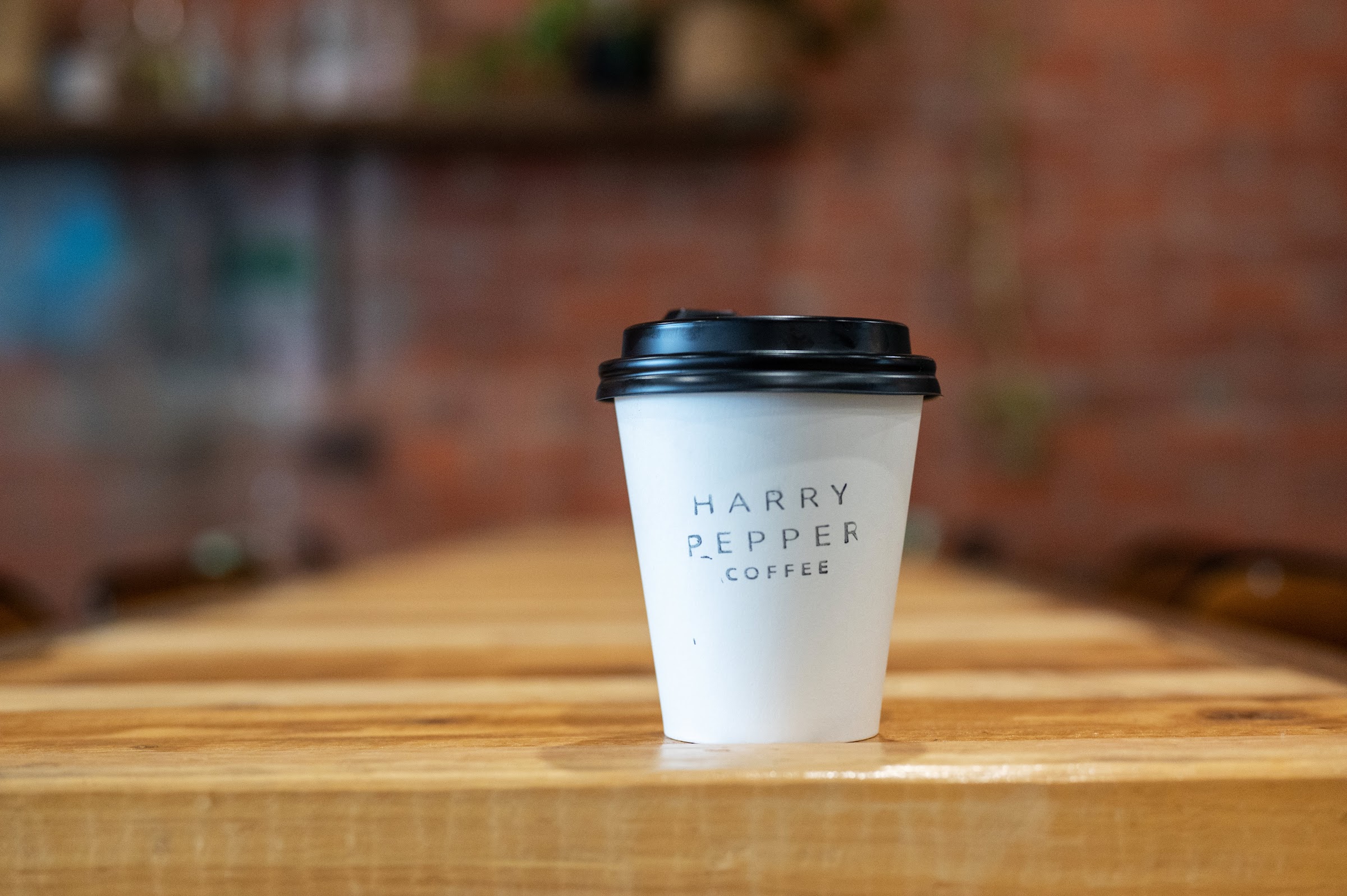 A Harry Pepper Coffee takeaway cup on a timber bench in front of the brick wall